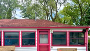 A cottage-style building with a red door, multiple windows, and a chimney. There are two wooden benches outside, one with a flower pot.