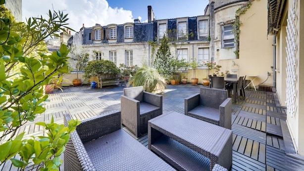 A courtyard with wicker furniture, potted plants, and a view of a building with multiple windows and a chimney. - Lovely  apartment with exceptional terrace (Bordeaux)