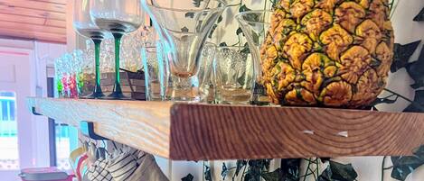 A bar area with a wooden shelf holding glasses and a pineapple. Below, there's a counter with a metal container, a mug, and a plate with items.