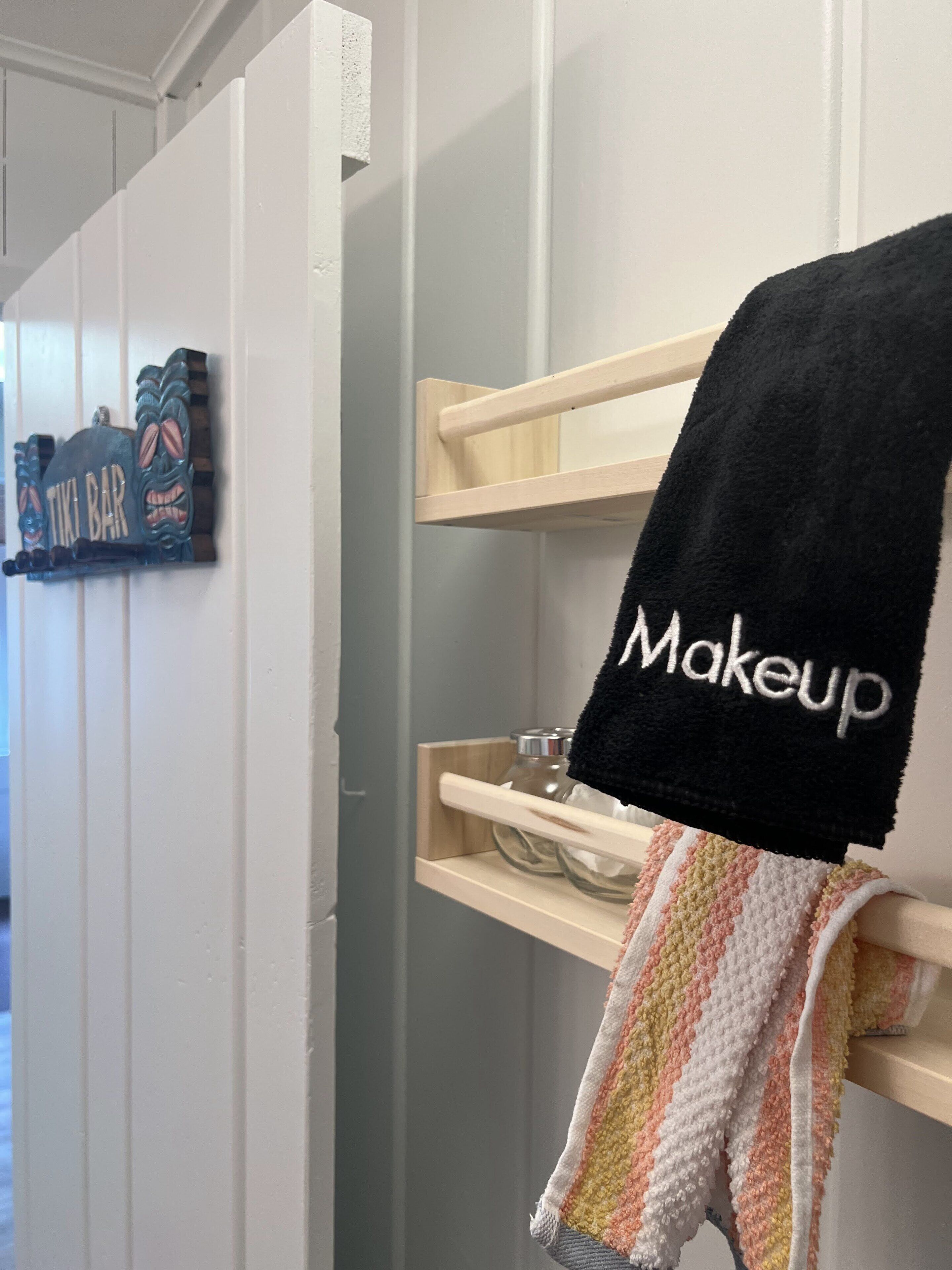 Un baño con un perchero que sostiene una toalla negra con el bordado 'Makeup', una toalla a rayas y un adorno con la etiqueta 'TIKI BAR'.