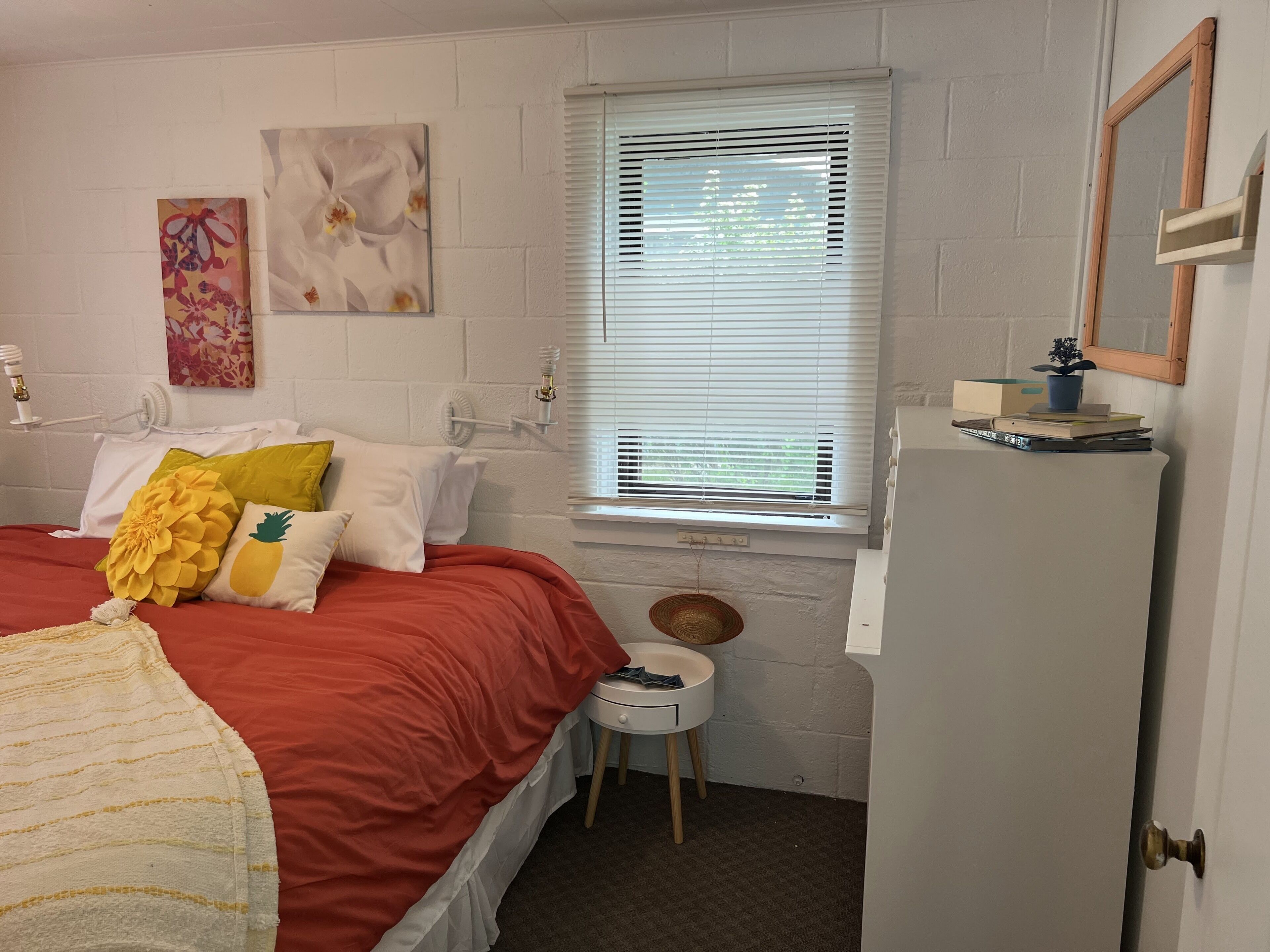 A bedroom with a bed, a window with blinds, a small table with a plant, and a door.