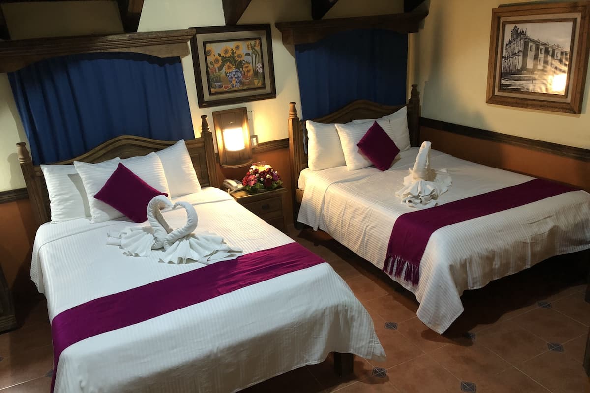 Deux lits bien faits, avec du linge de lit blanc et des touches de couleur violette, une tête de lit en bois et une table de chevet avec une lampe et des fleurs.