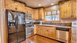 A kitchen with wooden cabinets, stainless steel appliances, and a window with blinds. - Gorgeous Forest Cabin with Hot Tub & Game Room (Greer)