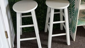 A bar area with a wooden countertop, two white stools, a glass of orange drink, a wooden chair, and a green leafy wall.