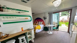 A hotel room with a bed, a desk with stools, a small table with a drink, and a view of the outside.