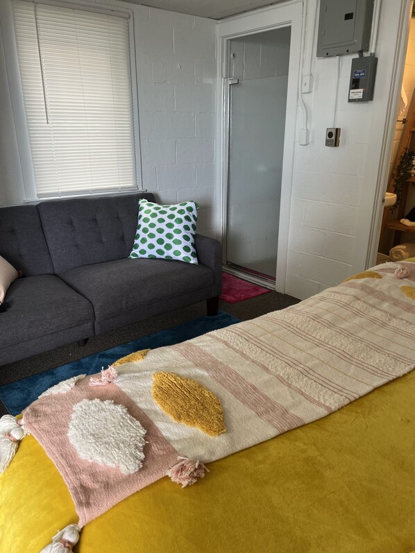 A room with a grey sofa, a patterned pillow, a large striped blanket, and a door with a light switch.