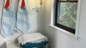 A corner room with a white bench, two wall-mounted towels, a shelf with baskets, and a window with a view of greenery.