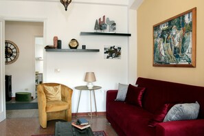 A living room with a red sofa, a yellow armchair, a small round table with a lamp, and a bookshelf with decorative items and books.
