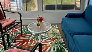 Quarto com sofá azul, mesa de vidro com garrafa e flores, e vista para o mar através de grandes janelas.