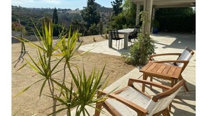Un patio al aire libre con mesa y sillas, rodeado de plantas y vistas a colinas.