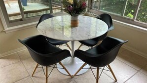 Una mesa redonda de vidrio con cuatro sillas negras, un florero con flores y vista a la vegetación a través de amplios ventanales.