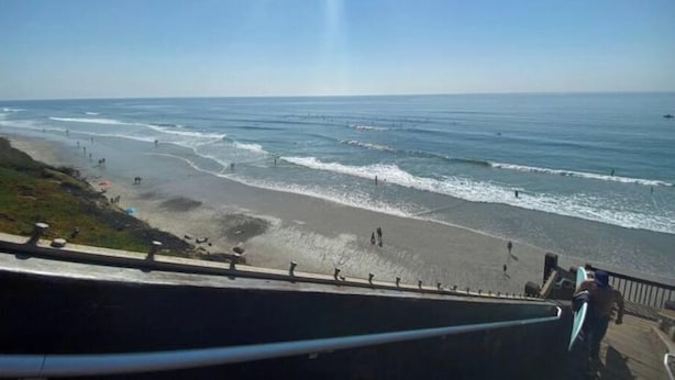 Una playa con olas, cielo despejado y una persona caminando.