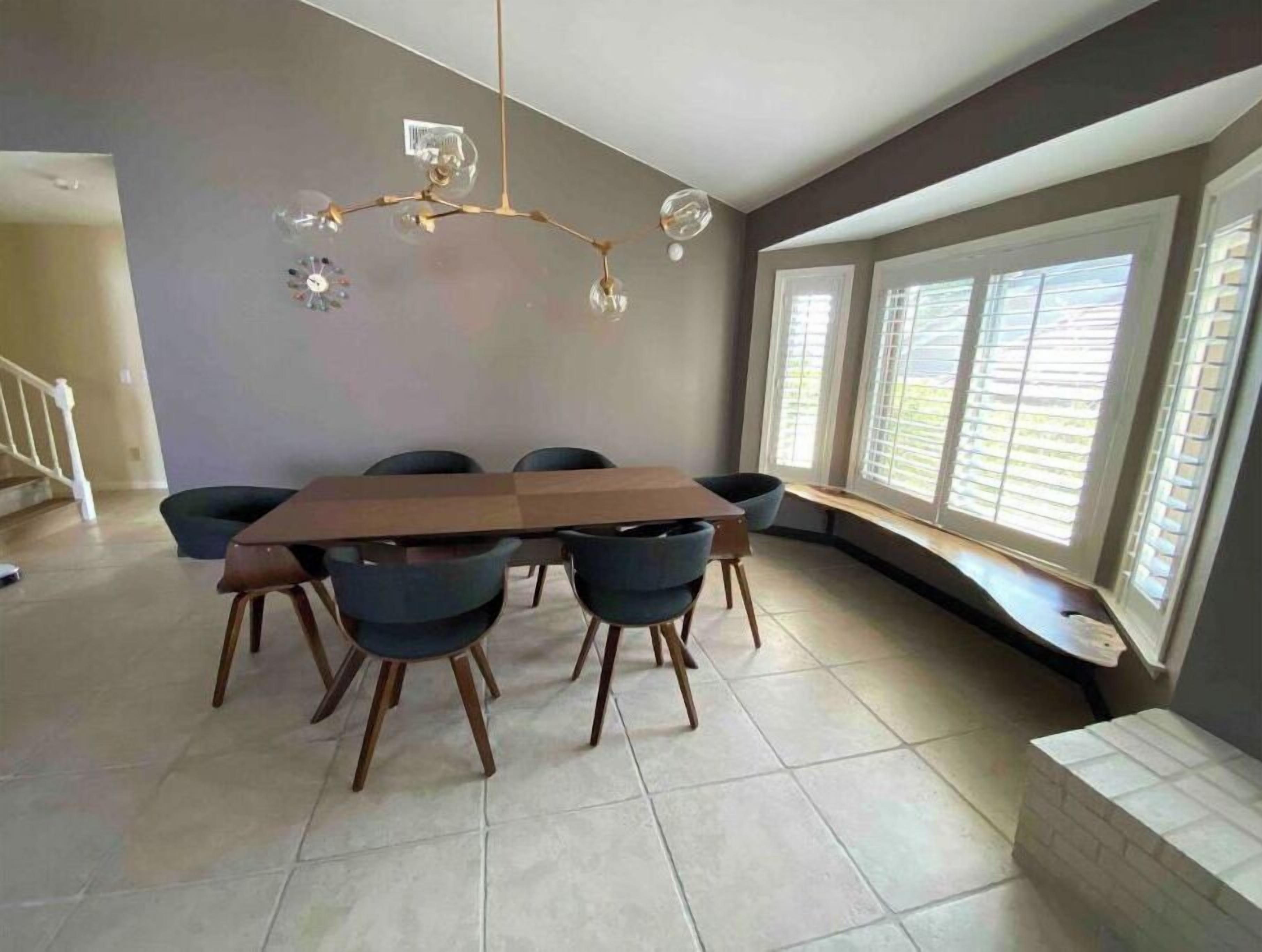 A dining area with a rectangular table surrounded by six chairs, a chandelier, and large windows with blinds.