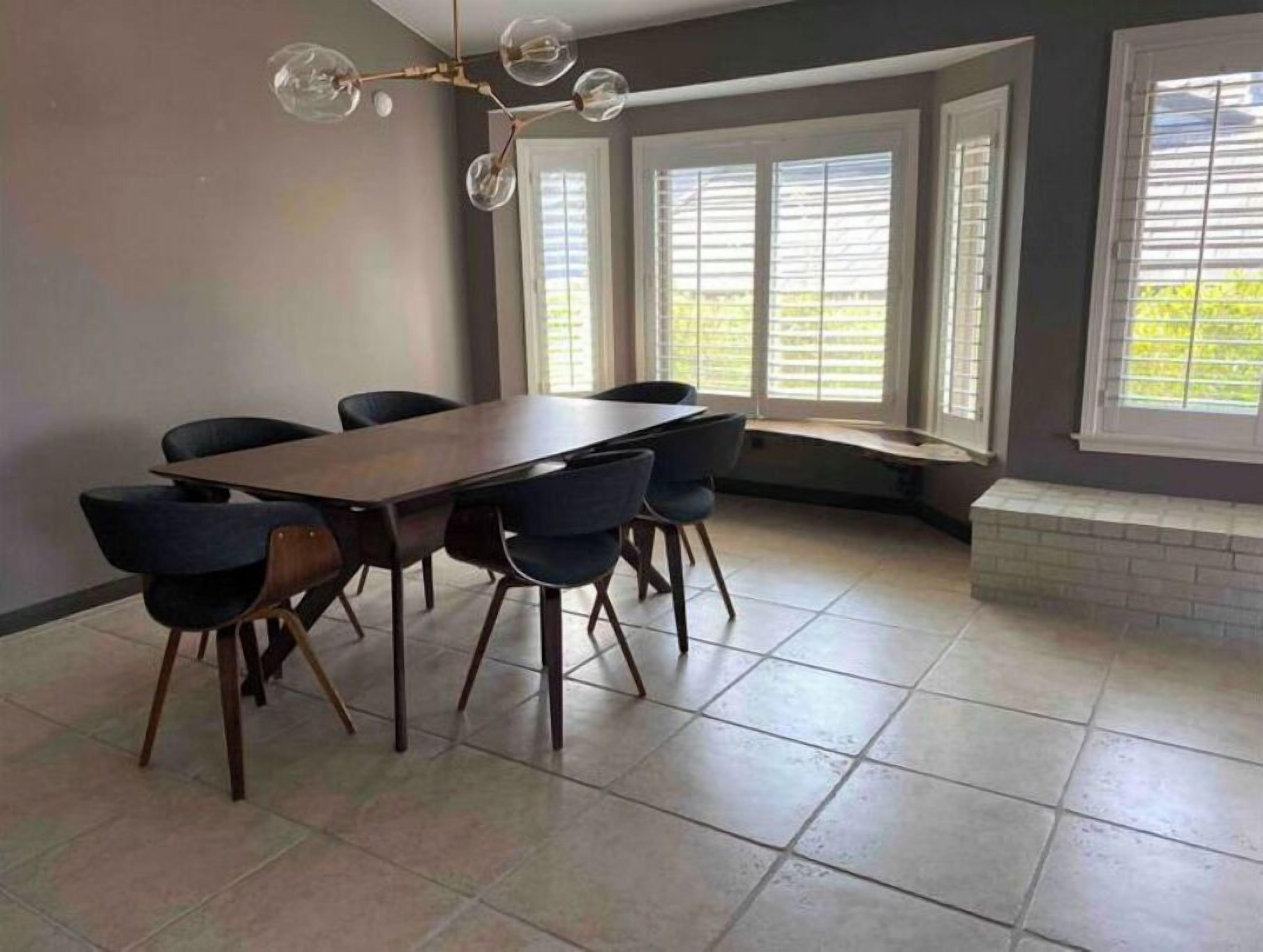 Un espace repas comprenant une longue table et six chaises, de grandes fenêtres équipées de stores et un lustre moderne.