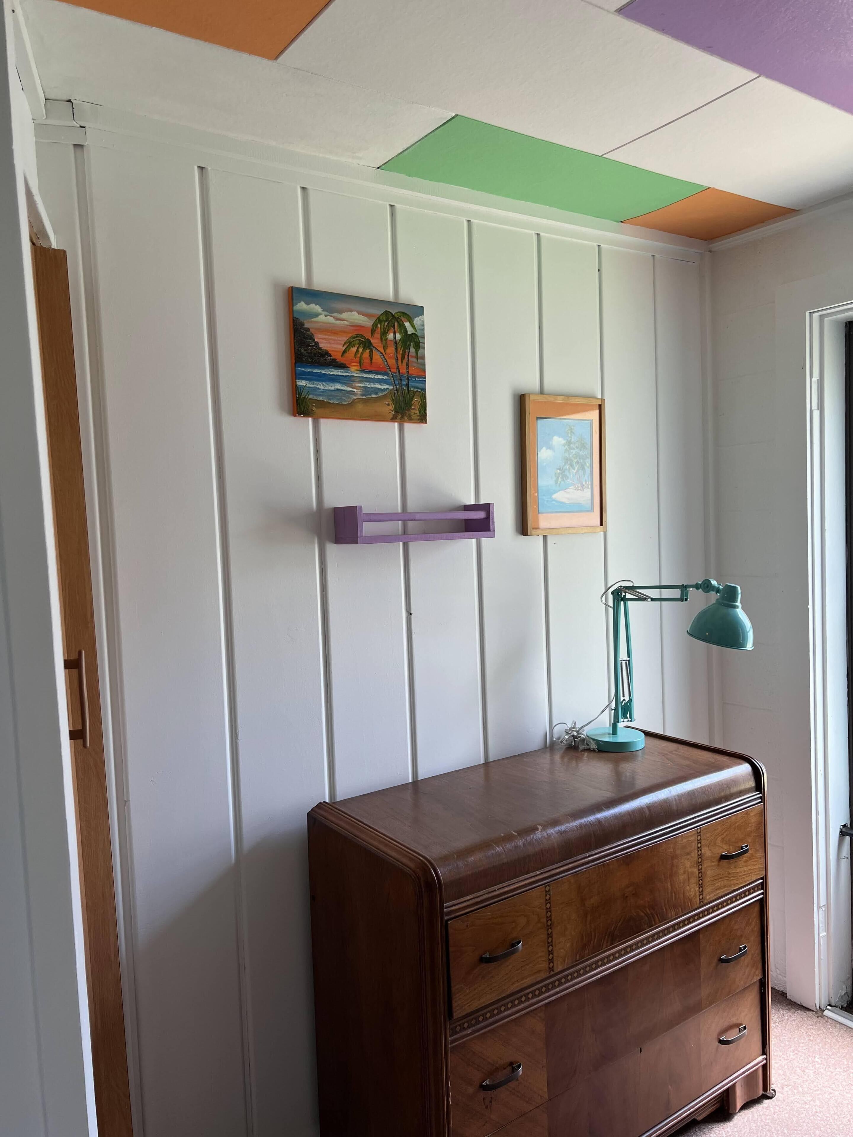 Una habitación con un aparador de madera, una lámpara de escritorio verde azulado y dos cuadros en una pared blanca con paneles.