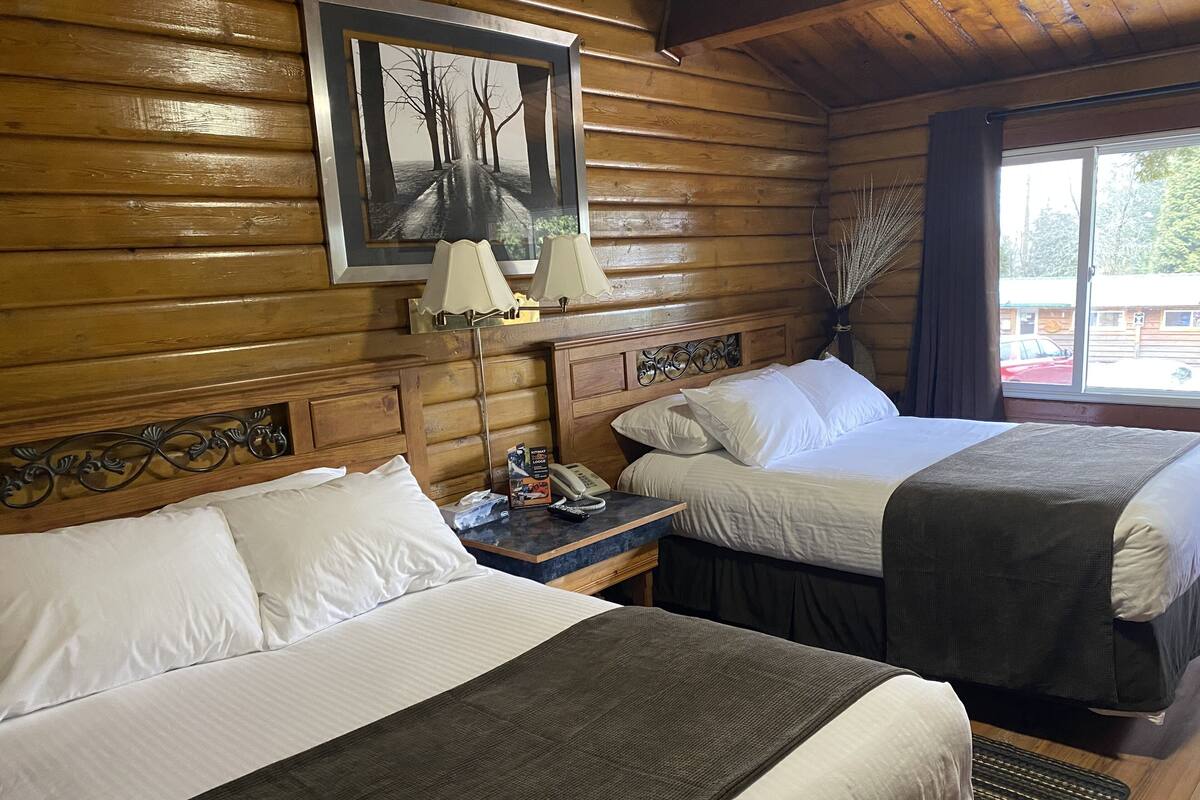 A wooden cabin room with two beds, a nightstand with a lamp, a framed picture on the wall, and a window with a view of the outside.