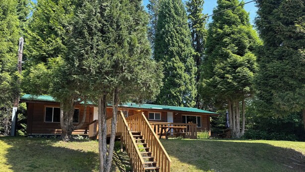 A cabin with a wooden staircase leading to a deck, surrounded by tall trees. - Kitimat Lodge (Kitimat)