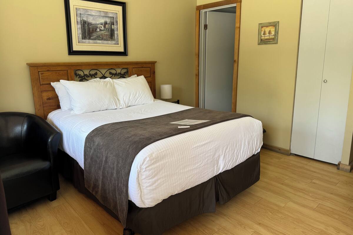 A neatly made bed with a wooden headboard, a black leather chair, a framed picture on the wall, and a wardrobe.