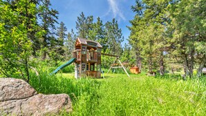 Une cabane en bois, avec un toboggan et des balançoires, installée dans un espace herbeux entouré d’arbres.