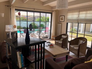 Uma sala de estar espaçosa com uma grande janela, vista para a piscina, móveis de vime e uma estante de livros.