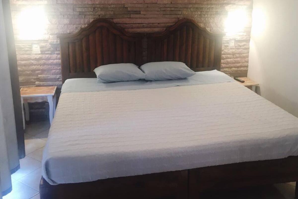 Una cama bien hecha con cabecera de madera, una mesita de noche pequeña y una mesita auxiliar.