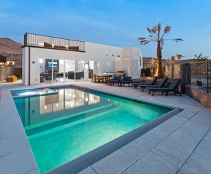 Et moderne udendørs poolområde med en klar, blå pool, et spiseområde med stole og bord samt et køkken med grill.