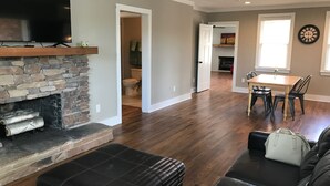 A spacious living room with a stone fireplace, a flat-screen TV mounted above it, a black leather ottoman, and a leather sofa. There is a dining area with a table and chairs visible in the background. - 1.3 Mile Walk to the Augusta National Clubhouse!<br>Lovely Cottage with Space! (Augusta)