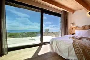 A bedroom with a large window, wooden ceiling beams, a bed with white linens, and a view of the landscape. - Casa Del Olivar (Aristotelis)