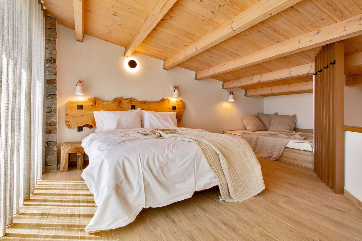 Un dormitorio con techo de madera, pared de piedra, cama con ropa de cama blanca y dos lámparas de noche.
