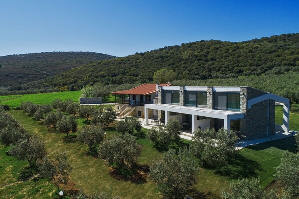 A modern house with a stone facade, large windows, and a red-tiled roof, surrounded by olive trees and a hillside. - Casa Del Olivar (Aristotelis)