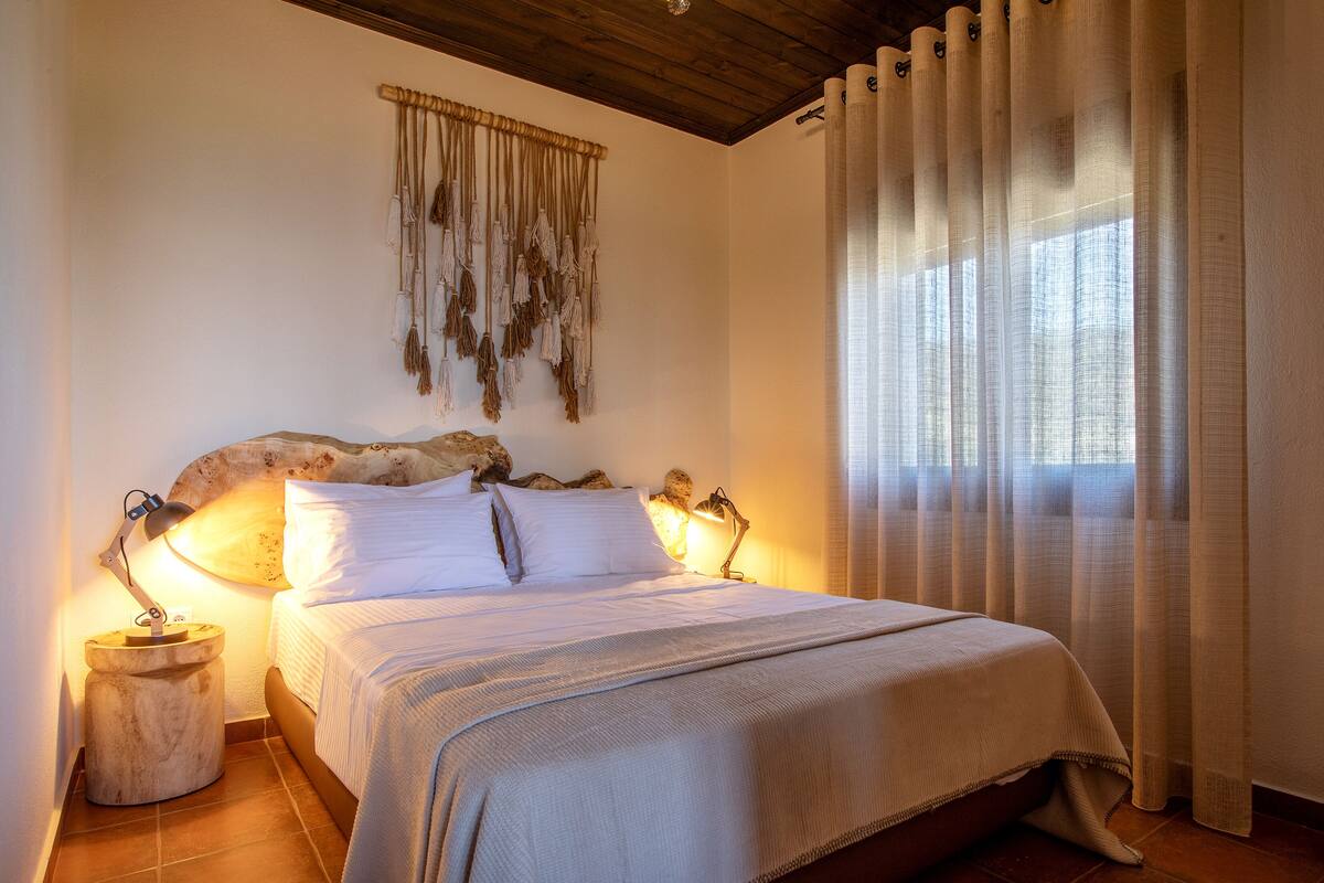 Una cama con ropa de cama blanca, dos lámparas de noche, un cabecero de madera, un adorno de macramé en la pared y cortinas de encaje.