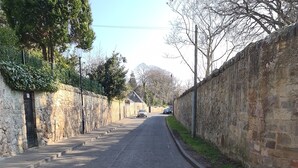 A narrow street with stone walls on both sides, a clear sky, and a few parked cars in the distance.