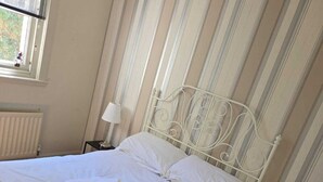 A neatly made bed with white linens and a rolled towel, a bedside lamp, and a window with blinds.