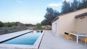 Una piscina con terraza de madera, una mesa con sillas y un edificio de techo con tejas.