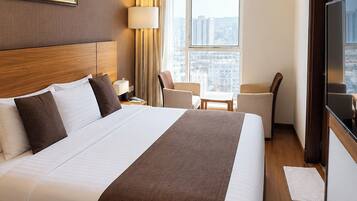 A hotel room with a large bed, a desk, a chair, a TV, and a window with a city view.