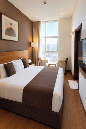 A hotel room with a large bed, a desk, a chair, a TV, and a window with a city view. - Nour Al Thuria Hotel (Makkah)