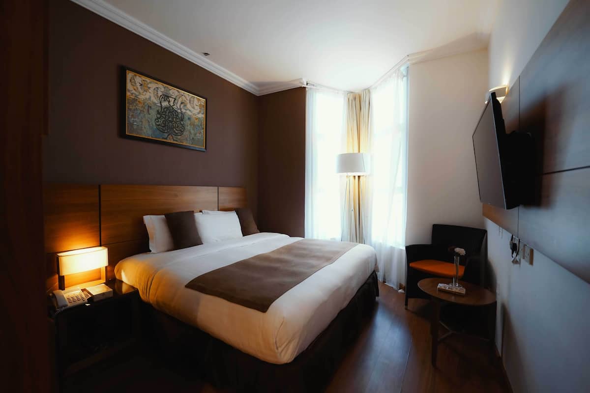Een hotelkamer met een groot bed, nachtkastje met een lamp, een klein tafeltje met een vaas, een stoel en een tv die aan de muur is bevestigd.