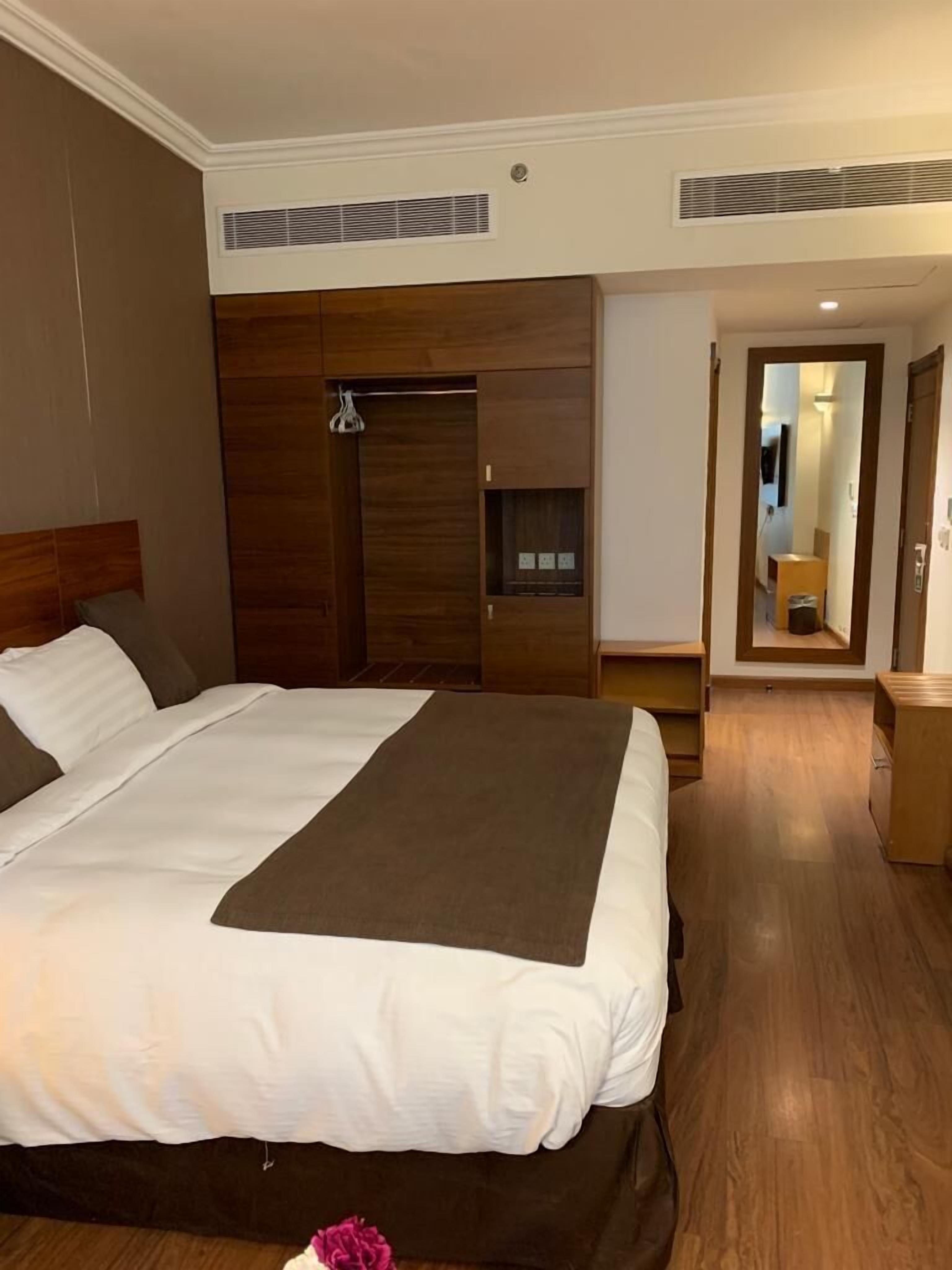 Une chambre d’hôtel dotée d’un grand lit, d’une tête de lit en bois, d’une armoire intégrée et d’un petit coin salon avec une table et une chaise.