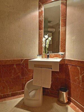 A bathroom with a marble wall, a sink, a mirror, a toilet, and a trash can. - Nour Al Thuria Hotel (Makkah)