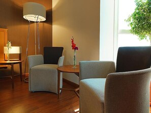 Een hotelkamer met een houten vloer, een ronde tafel met een bloevas, twee fauteuils, een vloerlamp en een raam met uitzicht op groen.
