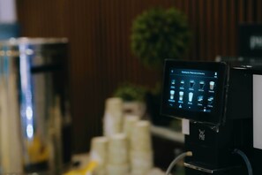 Een koffieapparaat met een digitaal menu, een stapel witte bekers en een plantje op de achtergrond.