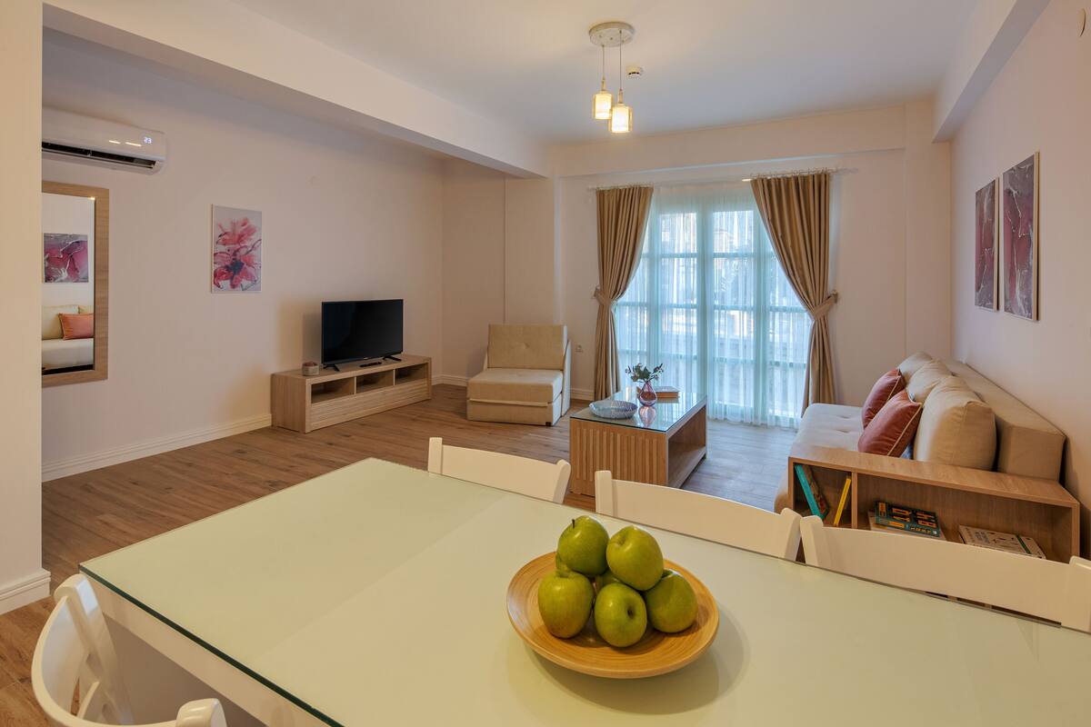 A modern living room with a glass dining table, a sofa, a TV, and a bowl of green apples on the table.