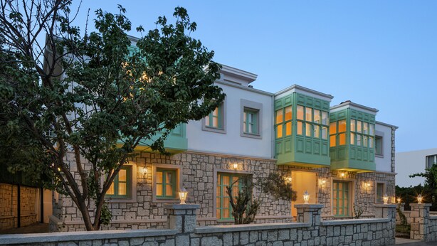 A two-story building with stone walls and green window frames, featuring multiple lit windows and a tree in the foreground. - Cigdem'inn Sigacik (Seferihisar)