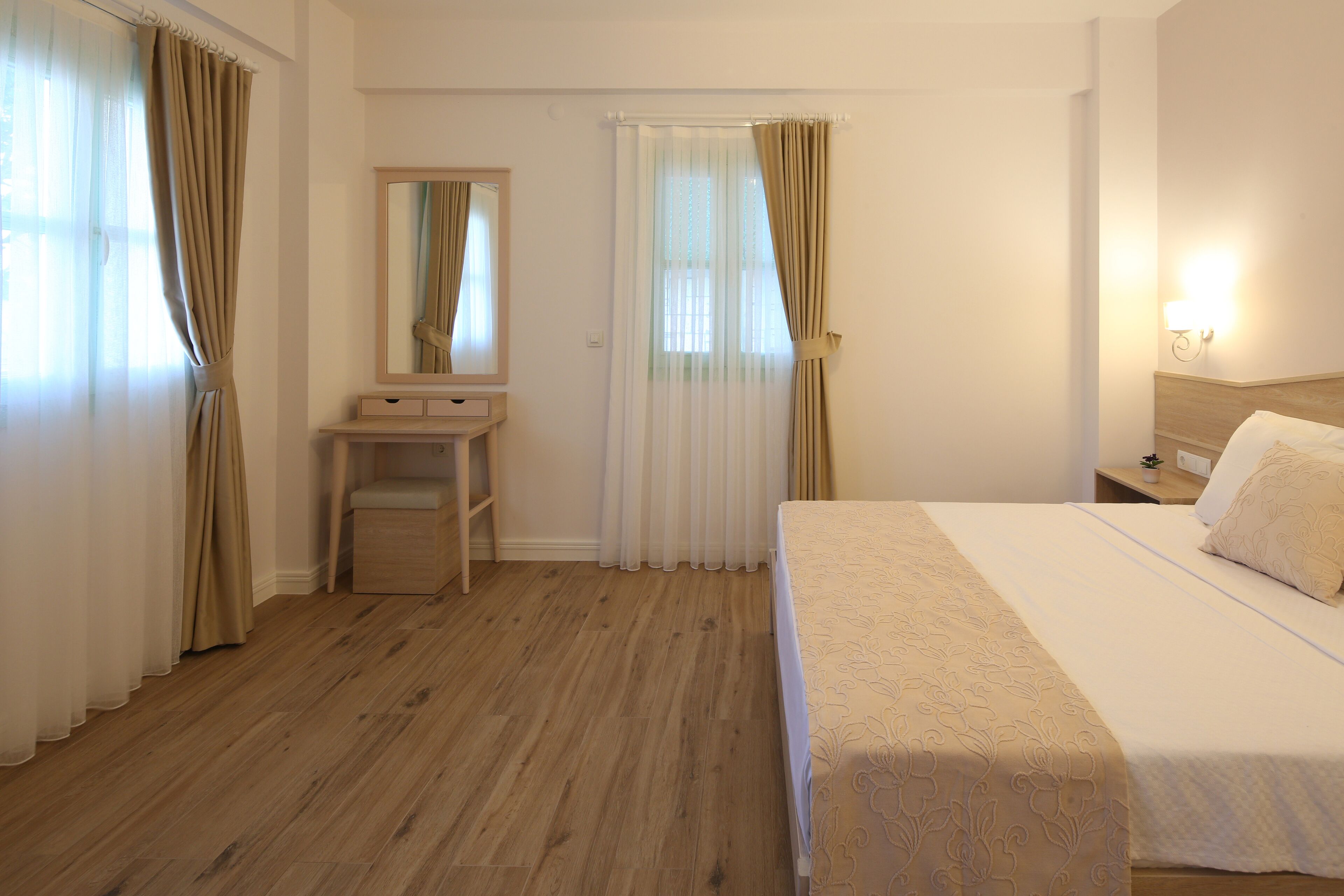 A hotel room with a wooden floor, a bed with a beige bedspread, a bedside table with a lamp, a mirror, and a window with curtains.