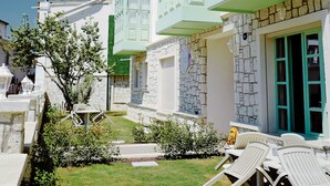 A building with a stone facade and green shutters, a small garden with a lawn and a few trees, and a seating area with white chairs. - Cigdem'inn Sigacik (Seferihisar)