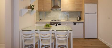 A compact kitchen with wooden cabinets, a gas stove, a sink, and a white refrigerator. There are four white chairs around a glass table with a bowl of fruit.