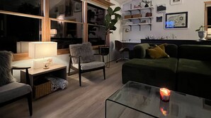 A modern living room with a glass coffee table, a sofa, a chair, and a desk with a lamp. There are shelves with decorative items and a large window with blinds. - Lake Superior premium bespoke accommodations with ultimate view! (Calumet Township)