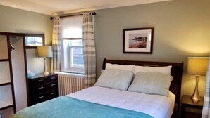 A neatly arranged bedroom with a bed, a bedside table, a lamp, a radiator, a window with curtains, and a framed picture on the wall. - Sunny Home B&B (Charlottetown)