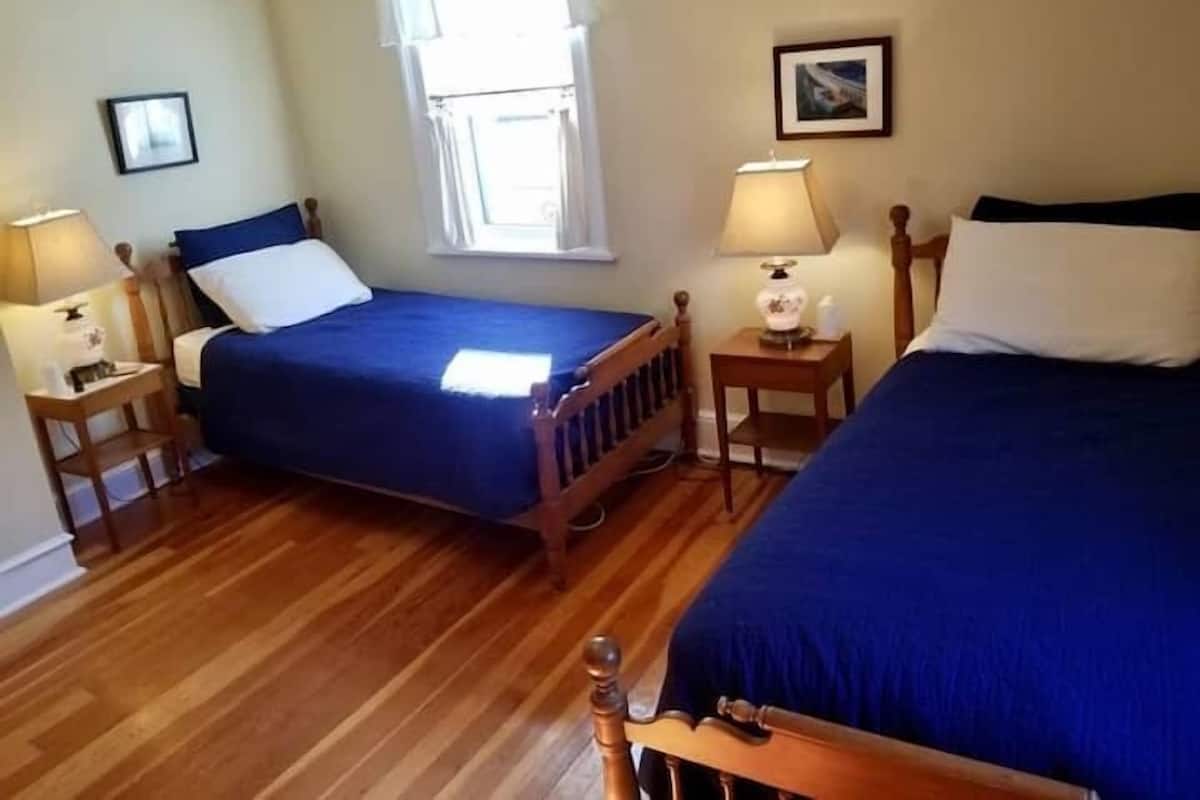 Een kamer met twee bedden, houten hoofdborden, een raam met gordijnen, twee nachtkastjes met lampen, en ingelijste foto's aan de muur.