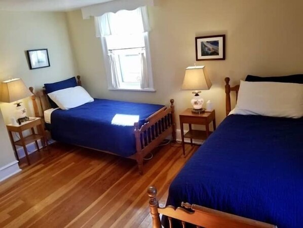 Una habitación con dos camas, cabeceros de madera, una ventana con cortinas, dos mesitas de noche con lámparas y cuadros enmarcados en las paredes.